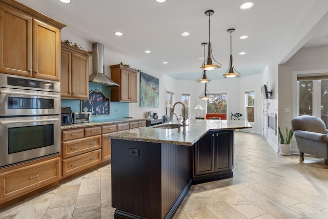 kitchen with hanging light fixtures, wall chimney exhaust hood, light stone countertops, an island with sink, and double oven