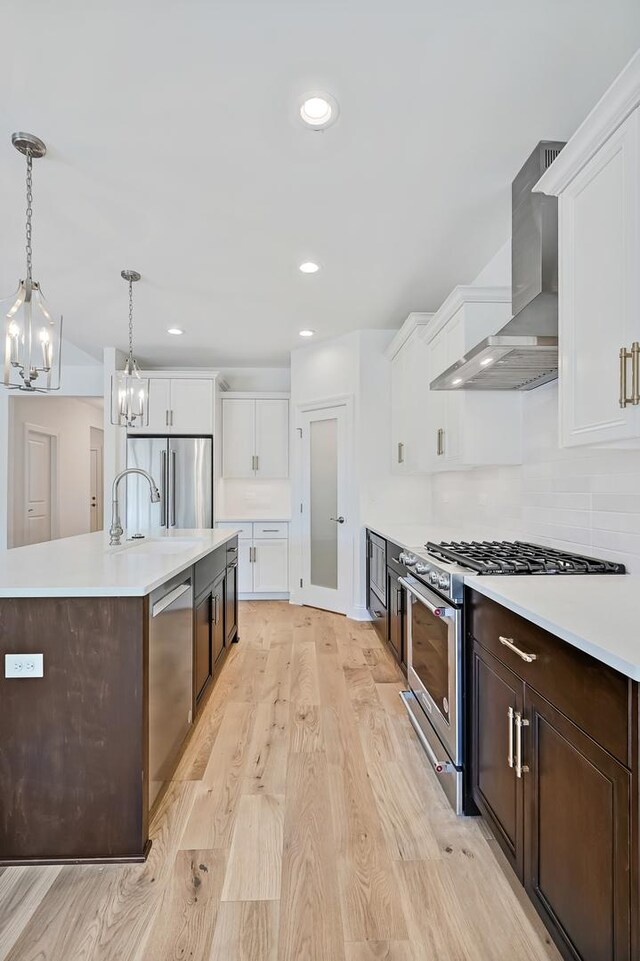 kitchen with wall chimney exhaust hood, light hardwood / wood-style flooring, an island with sink, stainless steel appliances, and white cabinetry