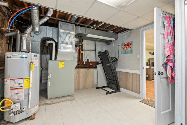 unfinished below grade area featuring heating unit, a drop ceiling, gas water heater, and wainscoting