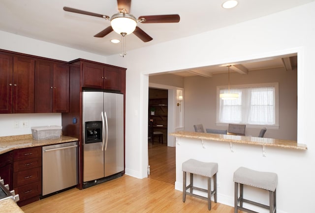 kitchen with appliances with stainless steel finishes, a breakfast bar, beamed ceiling, light stone counters, and light hardwood / wood-style flooring