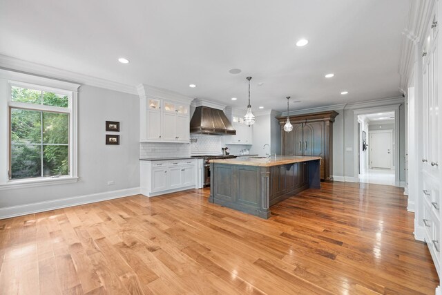 kitchen featuring white cabinets, decorative light fixtures, light wood-type flooring, wall chimney exhaust hood, and an island with sink