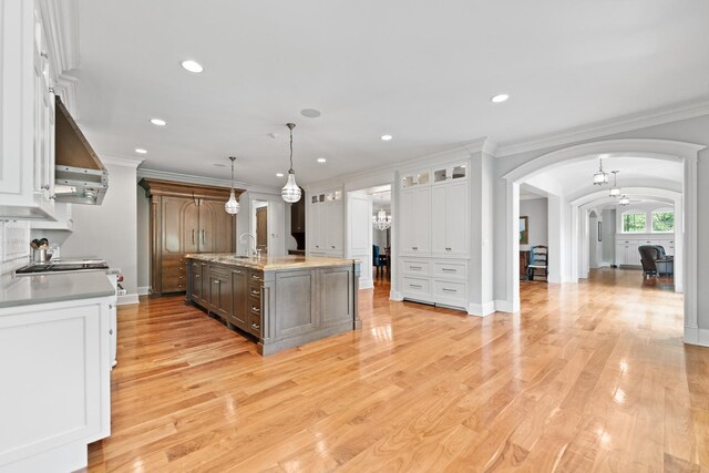 kitchen featuring a center island with sink, range hood, ornamental molding, white cabinetry, and light wood-type flooring