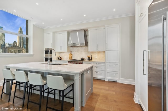 kitchen with white cabinets, wall chimney range hood, a kitchen island with sink, and high quality fridge