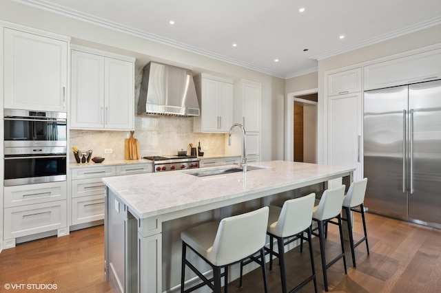 kitchen with appliances with stainless steel finishes, white cabinets, an island with sink, and wall chimney range hood