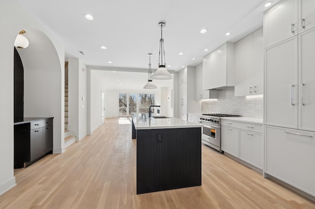 kitchen with stainless steel range, premium range hood, a center island with sink, white cabinets, and light wood-type flooring