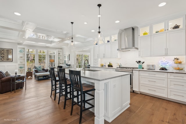 kitchen featuring an island with sink, stainless steel gas stove, wall chimney range hood, and white cabinetry