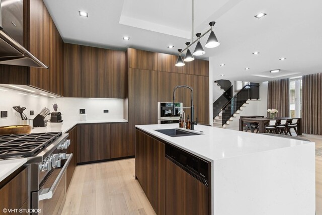 kitchen with a kitchen island with sink, light wood-type flooring, pendant lighting, stainless steel appliances, and wall chimney exhaust hood