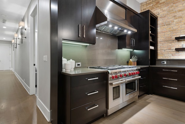 kitchen with tasteful backsplash, brick wall, light hardwood / wood-style flooring, wall chimney range hood, and range with two ovens