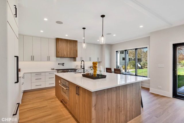 kitchen with an island with sink, light wood-type flooring, hanging light fixtures, and white cabinetry