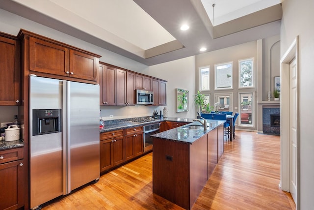 kitchen featuring light wood-type flooring, a kitchen island with sink, sink, appliances with stainless steel finishes, and dark stone countertops