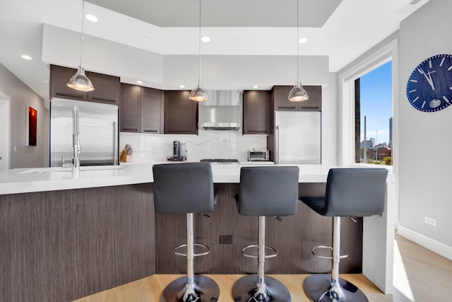 kitchen featuring hanging light fixtures, stainless steel fridge, wall chimney exhaust hood, built in fridge, and dark brown cabinets