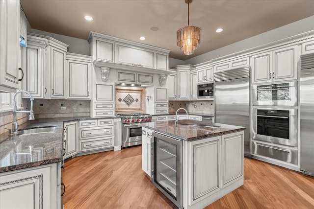 kitchen featuring built in appliances, wine cooler, dark stone countertops, an island with sink, and pendant lighting