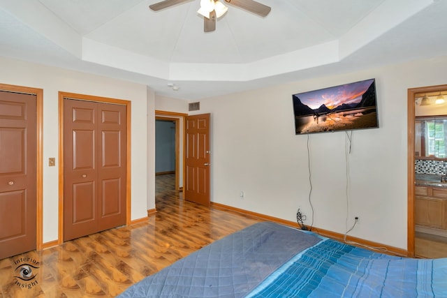 bedroom with light hardwood / wood-style floors, a tray ceiling, ceiling fan, two closets, and ensuite bath