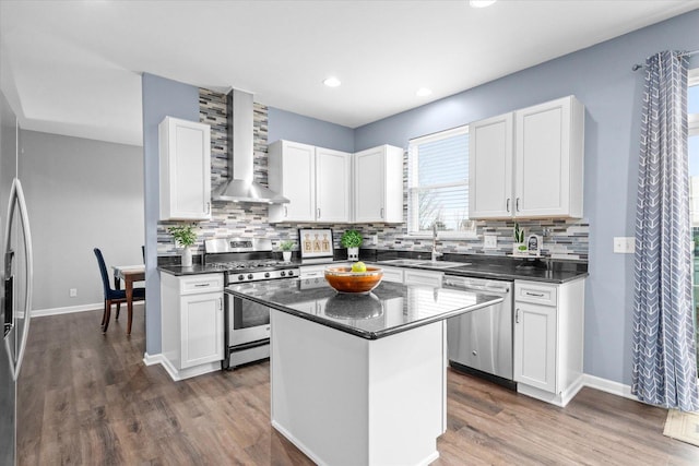 kitchen featuring wall chimney range hood, sink, appliances with stainless steel finishes, white cabinetry, and a kitchen island