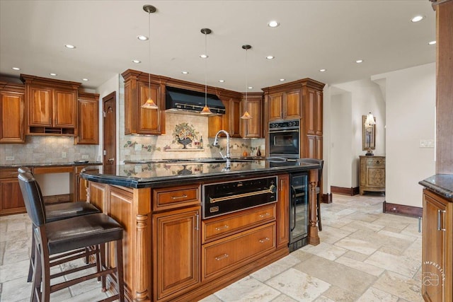 kitchen with hanging light fixtures, a kitchen breakfast bar, range hood, an island with sink, and oven