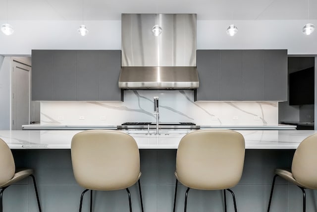 kitchen featuring gray cabinets, light stone countertops, wall chimney range hood, and a breakfast bar