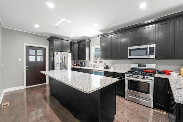 kitchen with stainless steel appliances, dark cabinets, a kitchen island, light stone counters, and wood finished floors