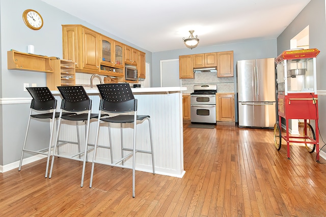 kitchen with light wood-type flooring, kitchen peninsula, a kitchen breakfast bar, backsplash, and appliances with stainless steel finishes