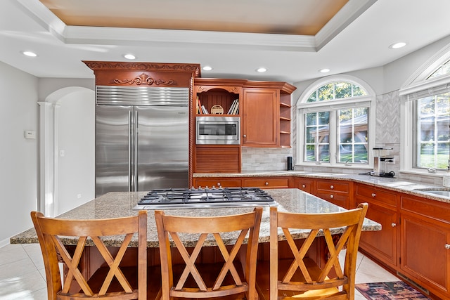 kitchen featuring light stone counters, tasteful backsplash, built in appliances, light tile patterned floors, and a tray ceiling