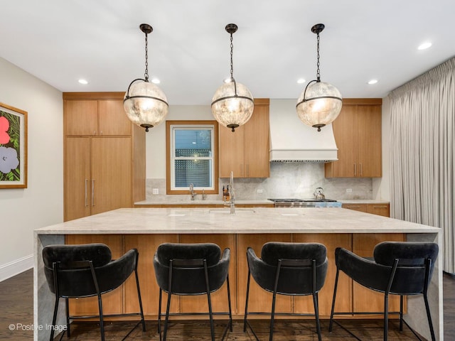 kitchen with premium range hood, tasteful backsplash, a sink, hanging light fixtures, and a large island with sink