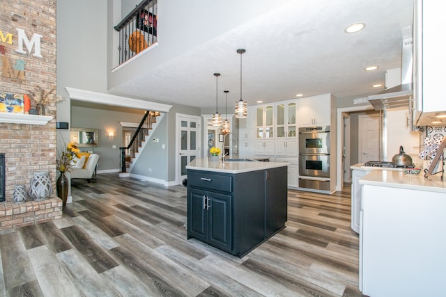 kitchen featuring an island with sink, a brick fireplace, white cabinetry, double oven, and hardwood / wood-style flooring