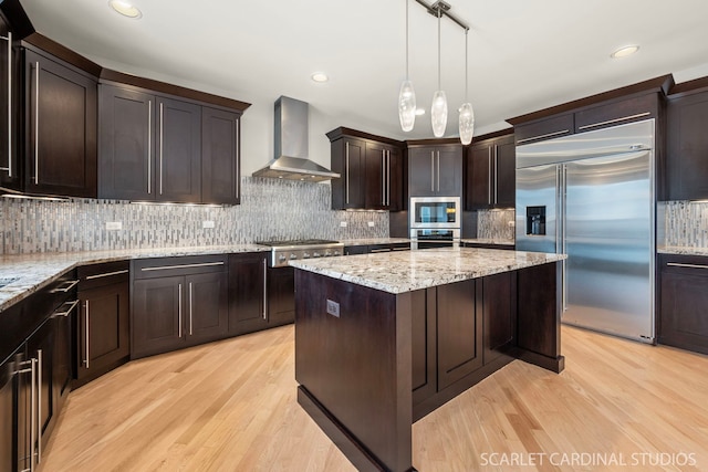 kitchen with wall chimney range hood, dark brown cabinets, built in appliances, light stone counters, and a kitchen island