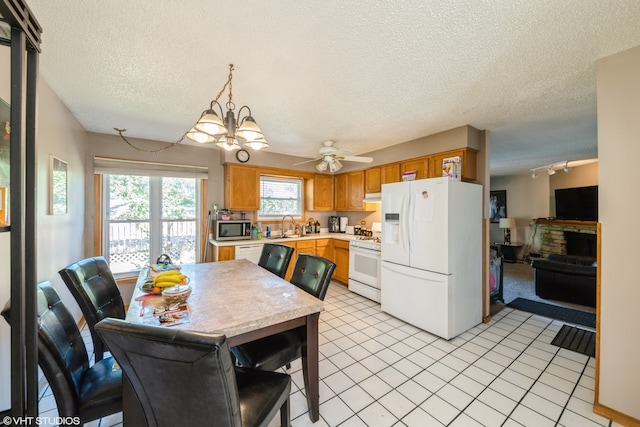 kitchen featuring a textured ceiling, ceiling fan with notable chandelier, white appliances, light tile patterned floors, and a stone fireplace