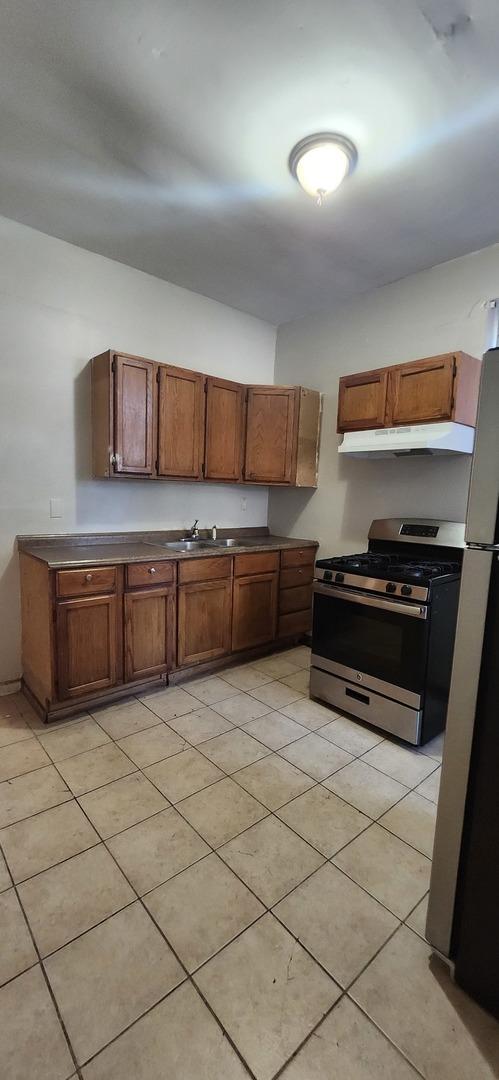 kitchen featuring brown cabinetry, dark countertops, freestanding refrigerator, under cabinet range hood, and gas stove