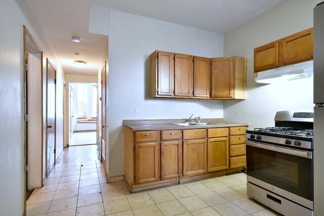 kitchen featuring light tile patterned floors, stainless steel gas range oven, under cabinet range hood, a sink, and brown cabinets