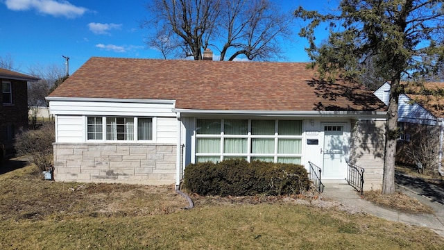 view of front of property featuring a front yard, stone siding, roof with shingles, and a chimney