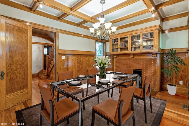 dining room featuring a brick fireplace, wood walls, light wood-type flooring, beam ceiling, and a notable chandelier