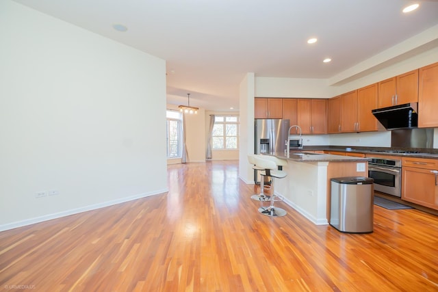 kitchen with stainless steel appliances, light hardwood / wood-style floors, hanging light fixtures, a breakfast bar area, and an island with sink