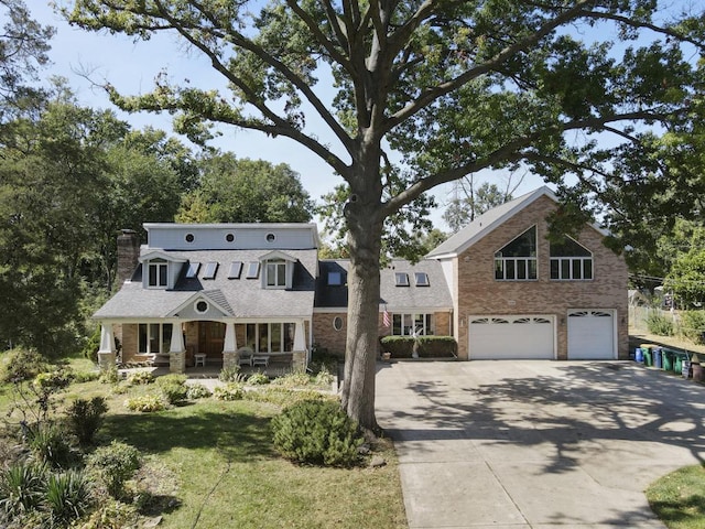 shingle-style home with a chimney, brick siding, an attached garage, driveway, and a porch