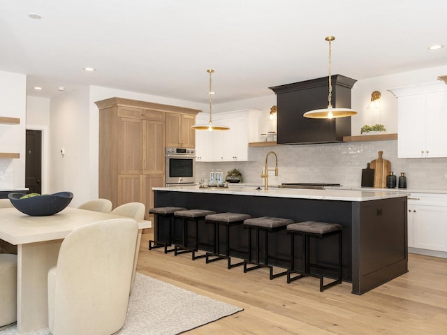 kitchen with white cabinetry, tasteful backsplash, hanging light fixtures, a kitchen island with sink, and oven