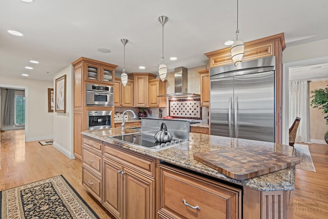 kitchen with wall chimney exhaust hood, light wood-type flooring, dark stone counters, stainless steel appliances, and a kitchen island with sink