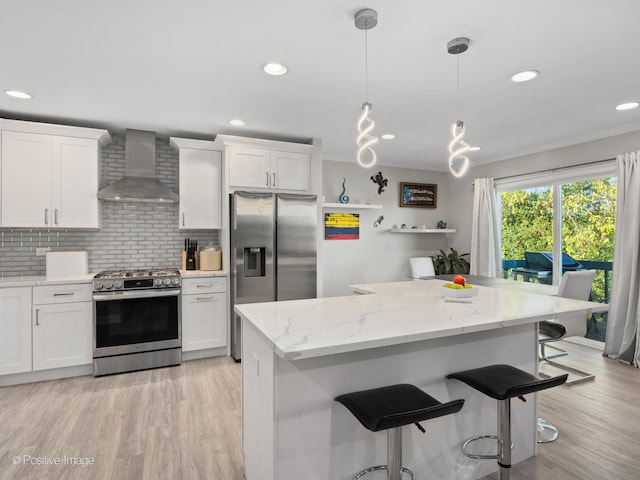 kitchen with a breakfast bar area, white cabinets, hanging light fixtures, wall chimney range hood, and stainless steel appliances