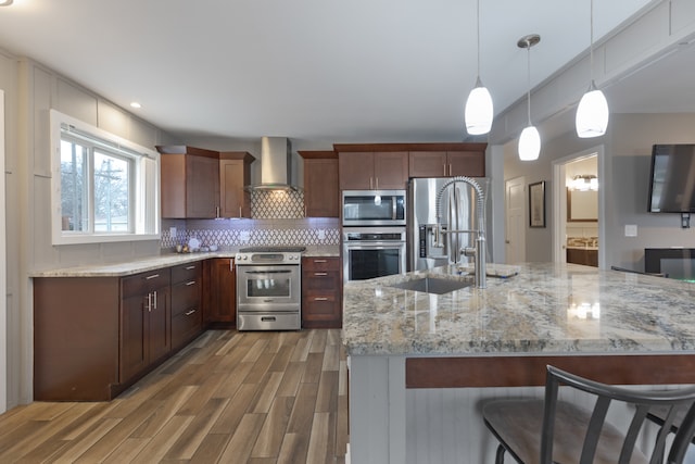 kitchen featuring wood-type flooring, decorative light fixtures, wall chimney range hood, appliances with stainless steel finishes, and a kitchen bar