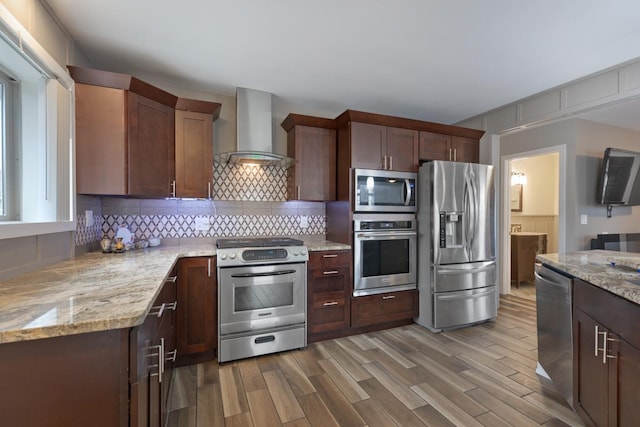 kitchen featuring tasteful backsplash, wall chimney exhaust hood, appliances with stainless steel finishes, wood finished floors, and light stone countertops