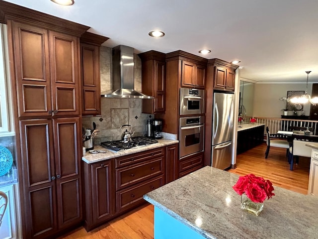 kitchen with light wood-type flooring, stainless steel appliances, crown molding, wall chimney range hood, and hanging light fixtures