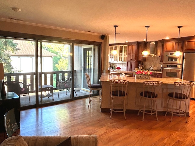 kitchen with decorative light fixtures, wall chimney exhaust hood, hardwood / wood-style flooring, stainless steel appliances, and a breakfast bar area