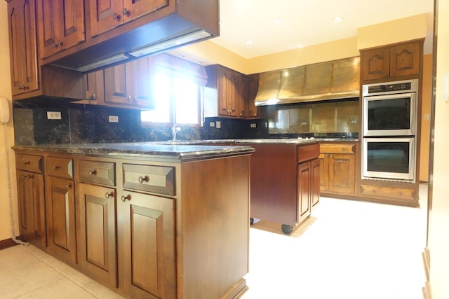 kitchen with double oven, light tile patterned flooring, backsplash, and wall chimney range hood