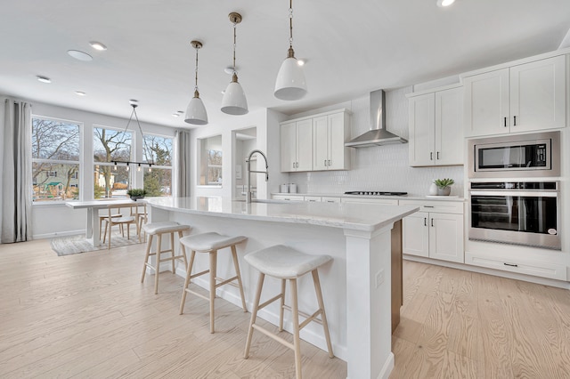 kitchen with white cabinets, a center island with sink, wall chimney exhaust hood, appliances with stainless steel finishes, and light wood-type flooring