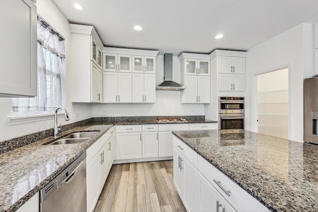 kitchen with sink, stainless steel appliances, wall chimney exhaust hood, dark stone counters, and white cabinets