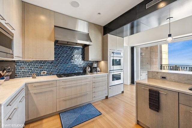 kitchen featuring wall chimney exhaust hood, backsplash, pendant lighting, light wood-type flooring, and appliances with stainless steel finishes