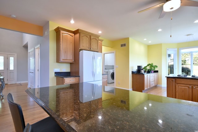 kitchen featuring ceiling fan, washer / dryer, dark stone countertops, light wood-type flooring, and white fridge