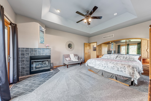 bedroom with dark colored carpet, a tiled fireplace, a raised ceiling, and ceiling fan