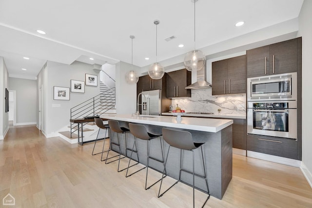 kitchen with light wood-style floors, light countertops, appliances with stainless steel finishes, wall chimney range hood, and an island with sink