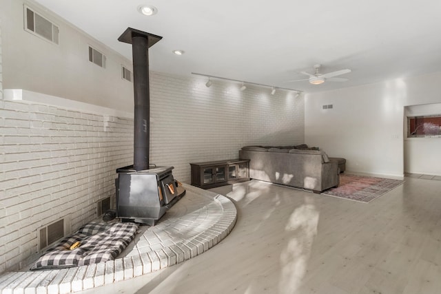 living room featuring a wood stove, ceiling fan, rail lighting, brick wall, and wood-type flooring