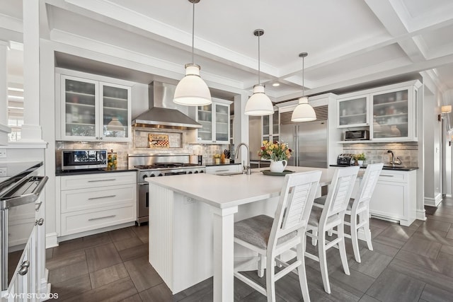 kitchen featuring beamed ceiling, hanging light fixtures, high end appliances, a kitchen island with sink, and wall chimney exhaust hood