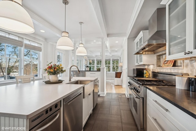 kitchen with wall chimney range hood, sink, appliances with stainless steel finishes, white cabinetry, and an island with sink